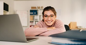 Junge Frau mit Brille lächelt in die Kamera und stützt sich mit verschränkten Armen auf einem Schreibtisch mit Laptop und Ordner ab. 