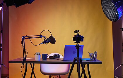 Ein Schreibtisch mit Laptop, Videokamera und Mikrofon steht vor einem orange ausgeleuchteten Screen. 