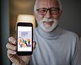 Ein weißhaariger Mann mit Bart und Brille hält lachend ein Smartphone in die Höhe. Auf diesem ist das Titelblatt der Broschüre "Erfolgreicher Einstige in die Nutzung von Smartphone und Tablet" zu sehen.