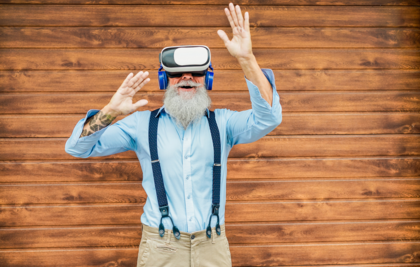 Ein Senior mit grauen Haaren und grauem Bart hat eine Virtual Reality Brille und blaue Kopfhörer an und hat Spaß