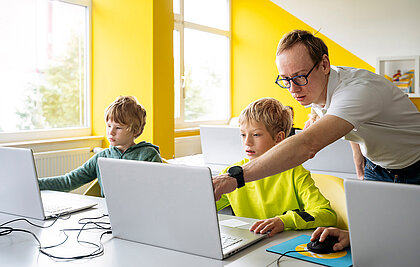 Lehrer in einer Grundschulklasse erklärt einem seiner Schüler das Arbeiten am Laptop.
