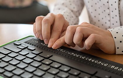 Die Hände einer Frau liegen einer Computertastatur mit Braillezeile und geben etwas in Brailleschrift ein.