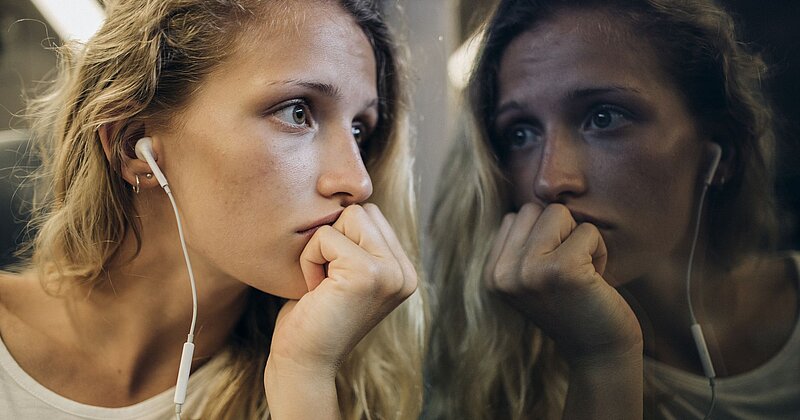 Ein Mädchen mit lockigen Haaren und Kopfhörern hält sich die Hand nachdenklich an den Mund und starrt aus dem Fenster einer U-Bahn, in dem sich ihr Spiegelbild zeigt. 