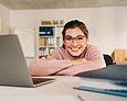 Junge Frau mit Brille lächelt in die Kamera und stützt sich mit verschränkten Armen auf einem Schreibtisch mit Laptop und Ordner ab. 