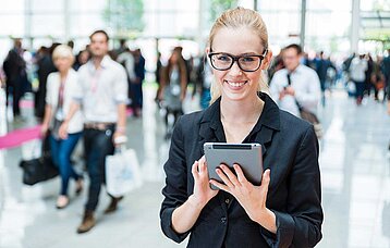 Freundlich, lächelnde Frau mit Tablet in der Hand. Im Hintergrund reges Messetreiben mit vielen Menschen in einer Messehalle.