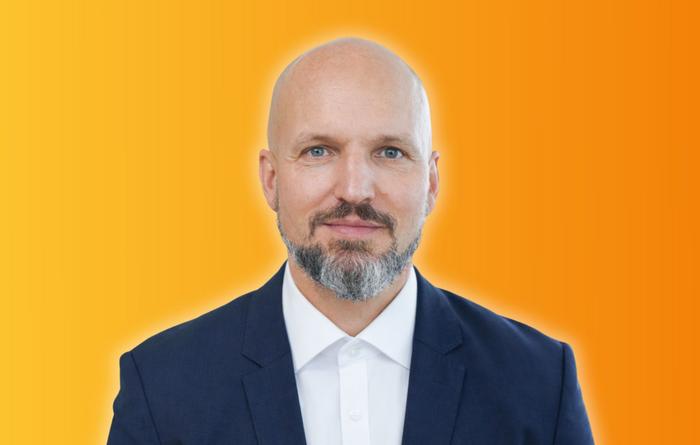 Mann mittleren Alters mit Glatze und grau-meliertem Bart in dunkelblauem Anzug und Hemd