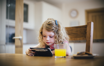 Mädchen im Grundschulalter sitzt am Tisch und schaut auf Smartphone