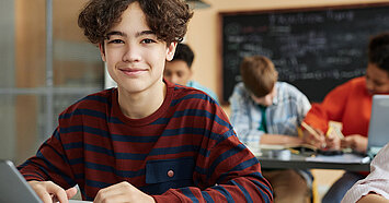 Schüler mit lockigem, braunen Haar lächelt in die Kamera. Er sitzt im Klassenzimmer und arbeitet am Laptop.