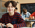 Schüler mit lockigem, braunen Haar lächelt in die Kamera. Er sitzt im Klassenzimmer und arbeitet am Laptop.