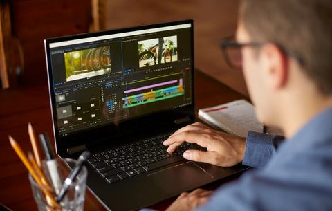 Ein Mann mit Brille ist an seinem Schreibtisch von hinten zu sehen. Er schaut auf einen Laptop, auf dem ein Videoschnitt-Programm geöffnet ist. 