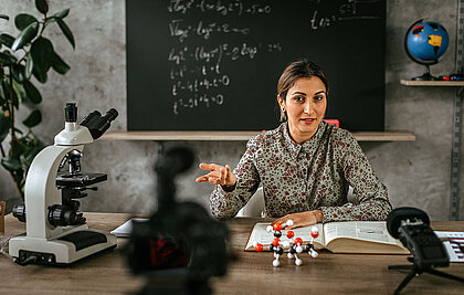 Lehrerin am Schreibtisch erklärt in einem Lernvideo Chemie. Im Hintergrund Tafel und Globus. 