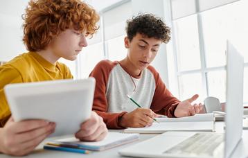Zwei Jugendliche unterhalten sich im Unterricht angeregt über eine Aufgabe. Sie arbeiten zusammen: Der Eine recherchiert im Tablet, der Andere notiert sich Ergebnisse. Beide arbeiten mit iPad und Tablet.