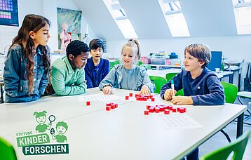 Fünf Schülerinnen und Schüler sitzen an einem Tisch und ordnen rote Würfel, die sie auf verschiedenen Blättern Papier anordnen. 