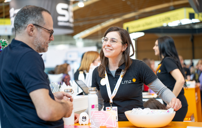 Junge Frau mit schwarzem LMZ-Polo-T-Shirt steht an einem Messestand und berät einen Messebesucher
