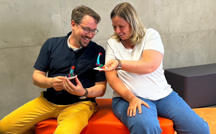 Zwei Lehrkräfte drehen sich sitzend zueinander und lachen beim Vergleich ihrer LEGO-Bauwerke.