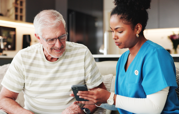 Ein alter Mann sitzt neben einer Pflegekraft, die gemeinsam mit ihm auf sein Handy schaut und über seine Elektronische Patientenakte spricht.  