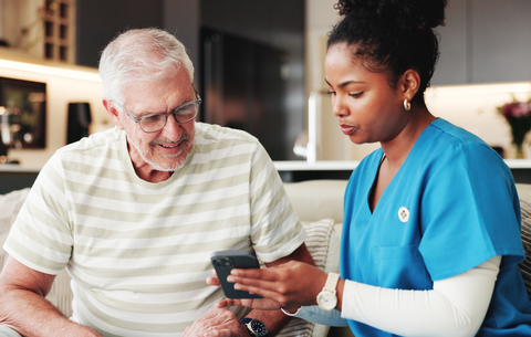 Gespräch über elektronische Patientenakte ePA Ein alter Mann sitzt neben einer Pflegekraft, die gemeinsam mit ihm auf sein Handy schaut und über seine Elektronische Patientenakte spricht.