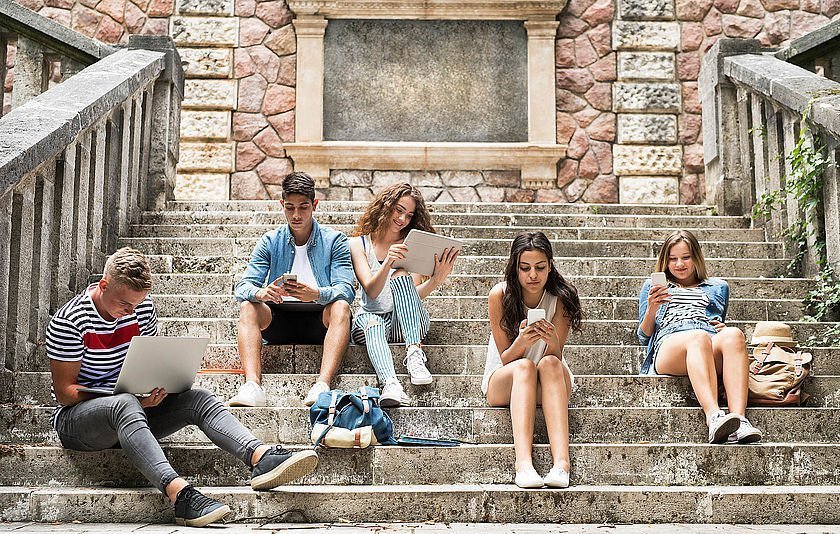 Schülerinnen und Schüler sintze auf einer antiken Treppe vor einer Schule oder Hochschule und schauen in ihre Handys und Tablets.