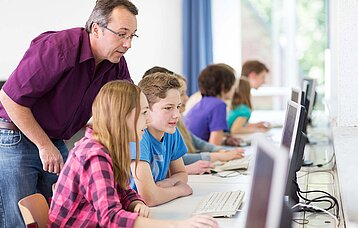 Ein Lehrer beugt sich von hinten über einen Schüler und eine Schülerin, um ihnen am PC etwas zuerklären. Die Atmosphäre wirkt vertrauensvoll.