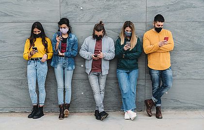 Jugendliche mit Maske lehnen an einer Mauer mit Blick in ihre Smartphones.