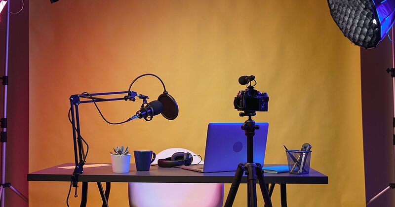 Ein Schreibtisch mit Laptop, Videokamera und Mikrofon steht vor einem orange ausgeleuchteten Screen. 