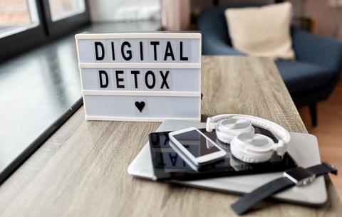 Ein Laptop, Tablet, Handy, Kopfhörer und eine Smartwatch liegen auf einem Schreibtisch. Dahinter steht ein Schild mit den Lettern "Digital Detox". 