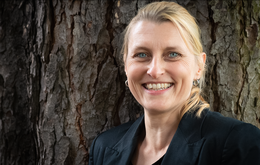 Frau mittleren Alters mit blondem Pferdeschwanz steht lachend vor einem Baum.