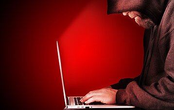 Ein Mann mit Kapuze sitzt gebeugt vor einem Laptop