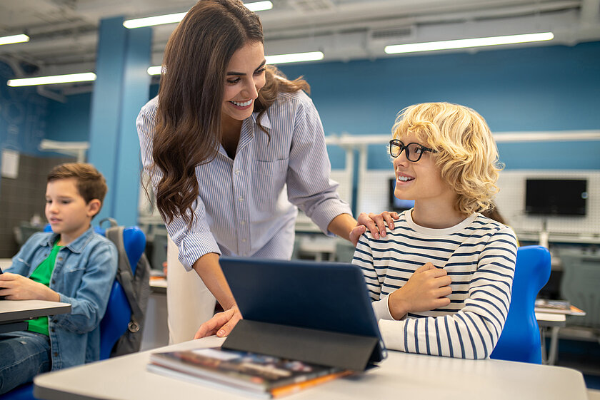 Lehrerin im Klassenzimmer mit Schüler. Beide lächeln. Auf dem Tisch steht ein Tablet.
