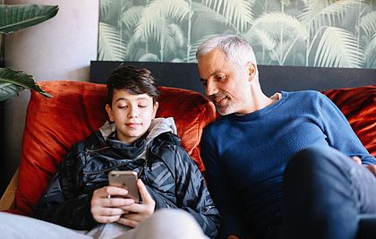 Ein junge und sein Vater sitzen auf dem Sofa. Der Junge zeigt seinem Vater etwas auf seinem Smartphone.