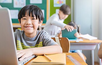 Kleiner, asiatischer Junge (Grundschüler) lächelt hinter seinem Laptop verschmitzt in die Kamera.