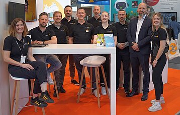 Zehn Personen stehen hinter einem Tresen versammelt am Messestand des Landesmedienzentrums auf orangenem Teppich.