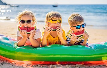 Drei Kinder im Grundschulalter liegen bäuchlings auf einer Luftmatratze am Strand. Ihre Gesichter sind durch Sonnenbrillen und Melonenschnitze vor dem Mund teilweise verdeckt.