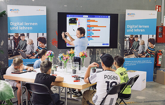 Fünf Jungen sitzen an Tisch mit Tablets und Kabeln. Lehrer steht im Hintergrund und zeigt Mini-Computer.