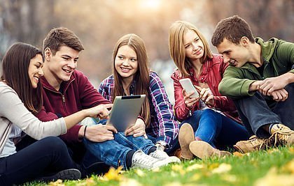 Jugendliche mit Tablets und Smartphones sitzen auf einer grünen Wiese.