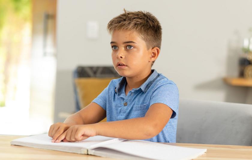 Ein Junge im Grundschulalter sitzt an einem Tisch, schaut in die Ferne und tastet mit seinen beiden Händen ein Buch in Braille-Schrift ab.