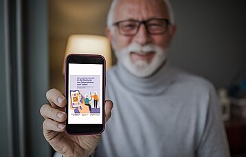 Ein weißhaariger Mann mit Bart und Brille hält lachend ein Smartphone in die Höhe. Auf diesem ist das Titelblatt der Broschüre "Erfolgreicher Einstige in die Nutzung von Smartphone und Tablet" zu sehen.