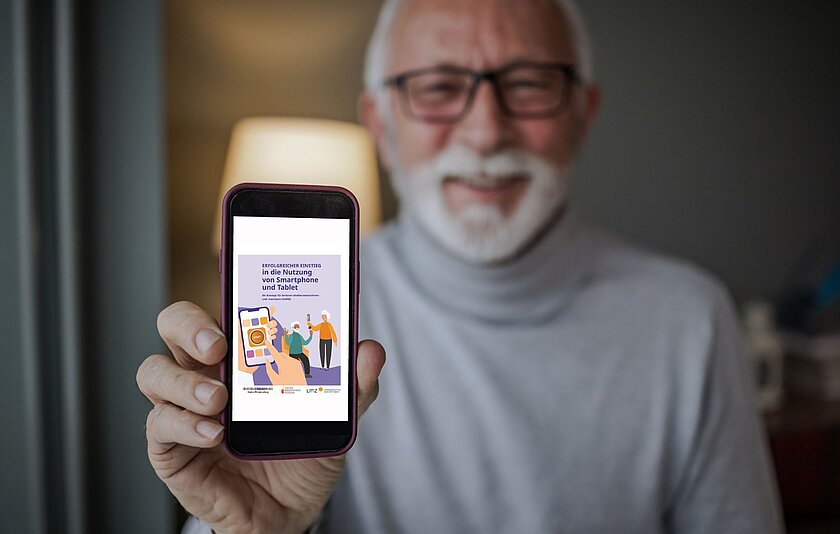 Erfolgreicher Einstieg in die Nutzung von Smartphone und Tablet Ein weißhaariger Mann mit Bart und Brille hält lachend ein Smartphone in die Höhe. Auf diesem ist das Titelblatt der Broschüre "Erfolgreicher Einstige in die Nutzung von Smartphone und Tablet" zu sehen.