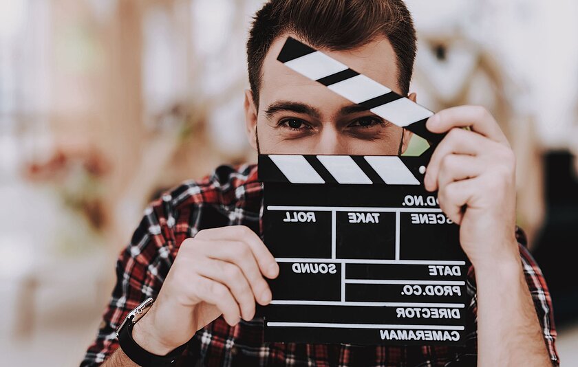 Ein Mann hält eine Filmklappe vor sein Gesicht, sodass nur noch die Augen erkennbar sind.