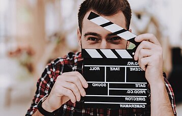 Ein Mann hält eine Filmklappe vor sein Gesicht, sodass nur noch die Augen erkennbar sind.