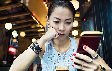 Eine junge Frau schaut ungläubig auf ihr Smartphone und hat ihre Hand nachdenklich auf ihr Kinn gestützt.