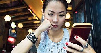 Eine junge Frau schaut ungläubig auf ihr Smartphone und hat ihre Hand nachdenklich auf ihr Kinn gestützt.
