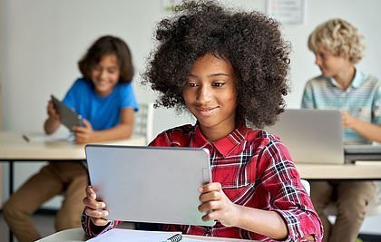 Afrikanisches Mädchen hält ein Tablet in der Hand und lächelt in den Monitor. Im Hintergrund sind zwei Schüler mit Tablets und lachen in der Klasse.