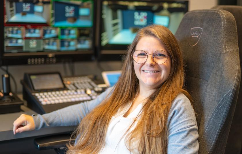 Barbara Glück vom LMZ Studio Eine junge Frau mit hellbraunen langen Haaren und runder Brille sitzt auf einem Regiestuhl. Hinter ihr sind das Regiepult und Monitore zu sehen.