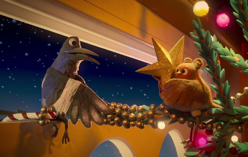 Ein Rabe und ein Rotkehlchen sitzen auf der Girlande eines Weihnachtsbaums. Das Rotkehlchen hält einen goldenen Stern in der Hand. 