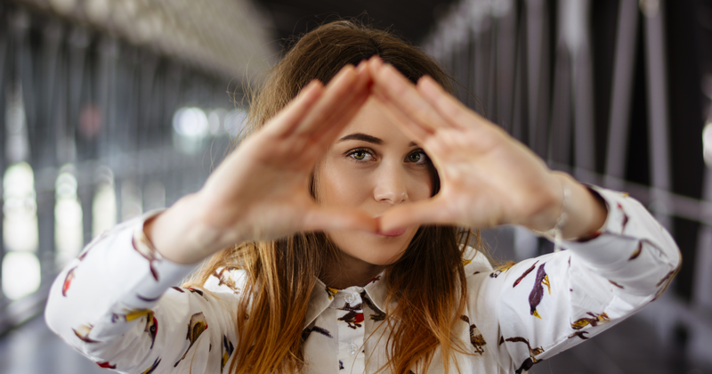 Eine junge Frau formt mit beiden Händen ein Dreieck und hält dieses vor ihr rechtes Auge.