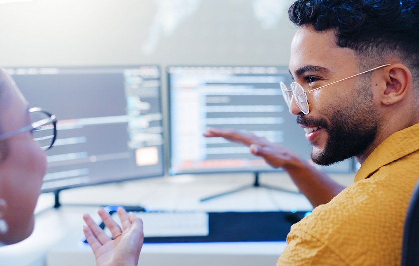 Blick von hinten auf zwei Personen, die sich freundlich miteinander fachlich vor ihren Bildschirmen austauschen.