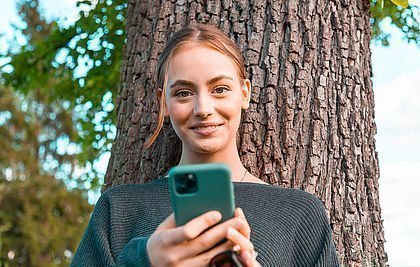 Glückliches Mädchen an einem Baum lehnend mit Handy in der Hand.