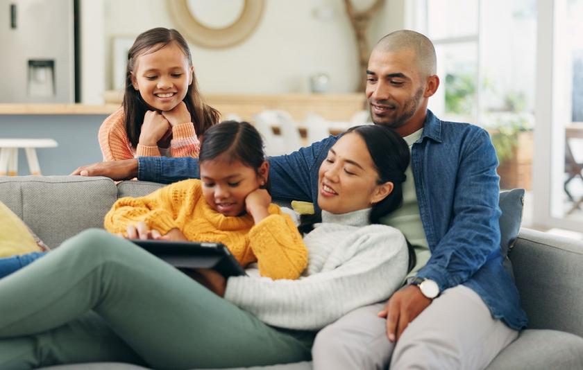 Medienerziehung in der Familie Eine vierköpfige Familie mit Vater, Mutter und zwei Töchtern sitzt gemeinsam gemütlich auf dem Sofa und schaut auf ein Tablet.