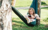Mädchen mit Tablet in der Hand liegt in einer Hängematte zwischen zwei Bäumen.